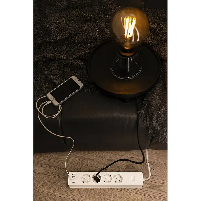 Osram Smart+ Smart-Steckdosenleiste 2. Gen. 2 USB-A, 2 USB-C - Weiß - Mehrfachsteckdose mit USB-Anschlüssen und einem Smartphone, das an einem Ladekabel angeschlossen ist, neben einer Glühbirne auf dem Sofa.