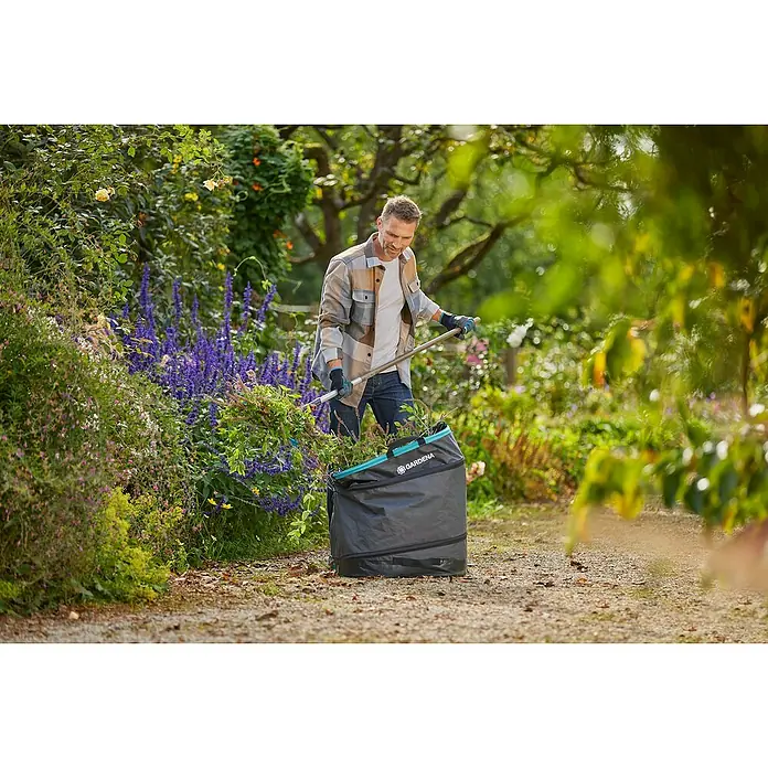 Gardena Pop-Up Gartenabfallsack Rechteck L - 220 l - Ein Mann füllt einen Gartensack von Gardena mit Pflanzenresten in einem üppigen Garten, während er Gartenarbeit macht.