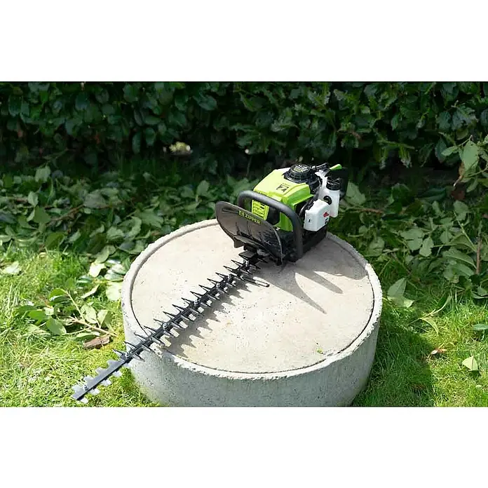 Zipper Benzin-Heckenschere ZI-BHS2600 - 700 W, Schnittlänge: 60 cm, Max. Aststärke: 27 mm - Nahaufnahme einer Heckenschere mit grünen Akzenten, die auf einem Betonsockel steht und von Grünschnitt umgeben ist.