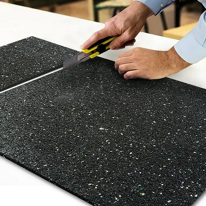 Floordirekt Unterlagsmatte Antivibrationsmatte 60 x 60 x 0,5 cm - 0,36 m², Stärke: 5 mm - Eine Person schneidet eine strapazierfähige Gummimatte mit einem gelben Cuttermesser. Ideal für Fitnessstudios oder Werkstätten.