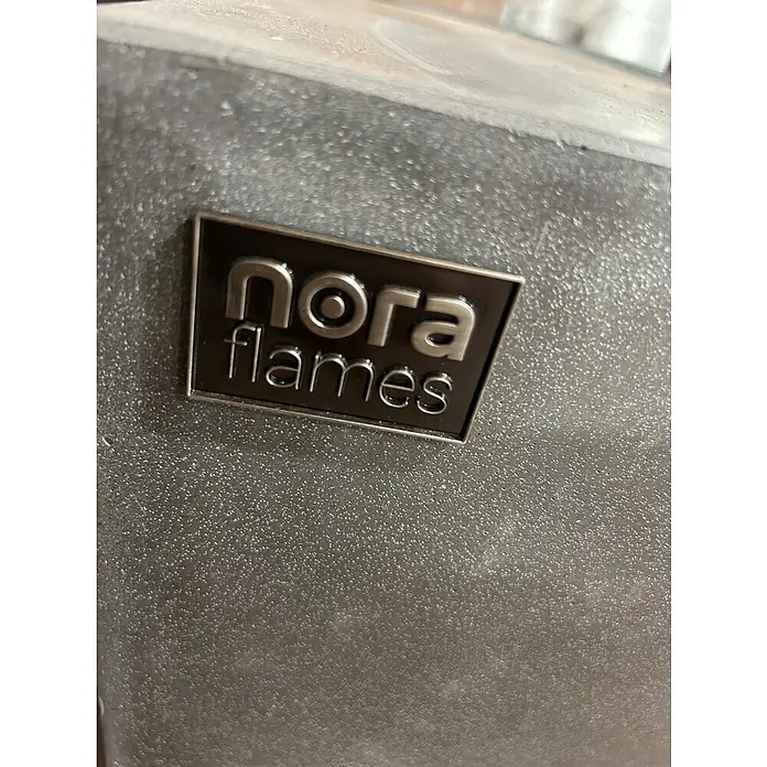 Nora Flames Estufa de exterior Colima - 16 kW, Altura: 40 cm, Gris - Primer plano de un logotipo de Nora Flames en un producto. El logo es plateado sobre un fondo oscuro y texturizado.