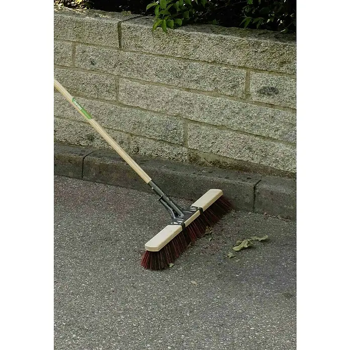 Freund Victoria Straßenbesen Profi - Arbeitsbreite: 60 cm, Material Borsten: Elaston, Natur, Schwarz/Rot - Bild einer Straßenbürste mit Holzstiel und roten Borsten, die auf einer grauen Asphaltfläche vor einer Steinmauer liegt.