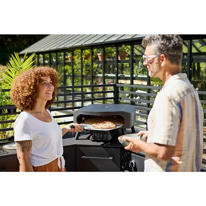 Cozze Pizzaofen Black Edition 17" mit drehbarem Pizzastein - Durchmesser Grillfläche: 43 cm, 8 kW - Eine Frau, die eine Pizza aus dem Lavastaingrill nimmt, während ein Mann eine Schüssel hält, alles im Freien.