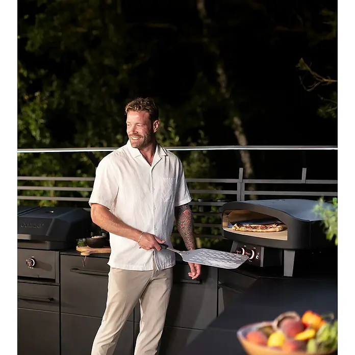 Cozze Pizzaofen Black Edition - Grillfläche (B x T): 60 x 60 cm - Ein Mann bereitet Pizza in einem Ooni Pizzaofen im Freien zu, der für seine schnelle Zubereitung und hohe Temperaturen bekannt ist.