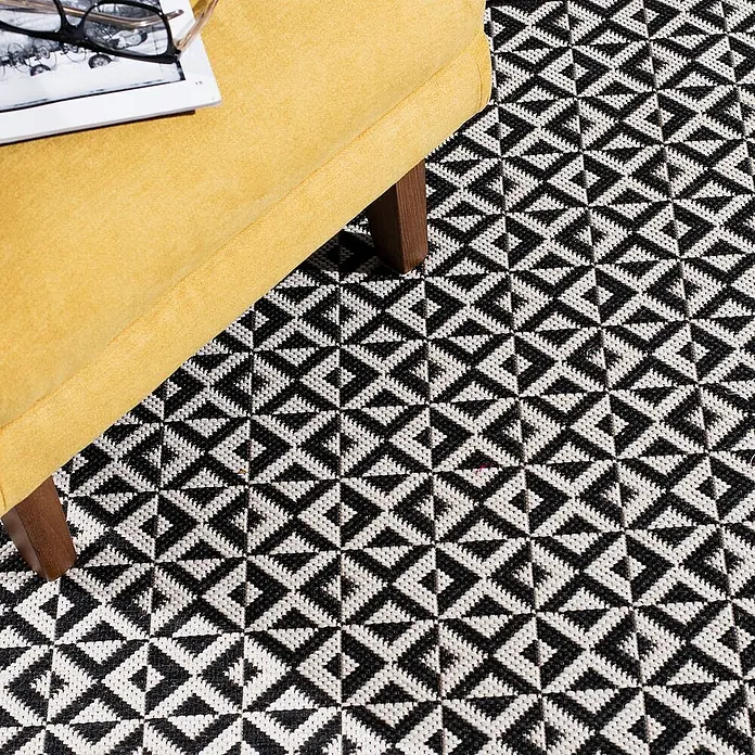 Dekoria Flachgewebeteppich - weiß, schwarz, 170 x 120 cm - Nahaufnahme eines modernen, schwarz-weißen geometrischen Teppichs unter einem gelben Sessel.