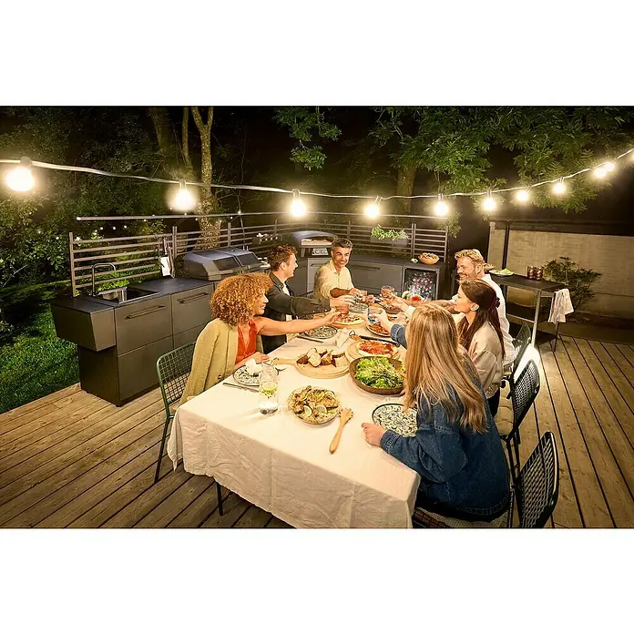 Cozze Hybrid-Grill Quattro 900 - Grillfläche (B x T): 72,7 x 34,5 cm, 13 kW, Anzahl Brenner: 3 Stk. - Gesellige Runde an einem Esstisch im Freien, beleuchtet von einer Lichterkette, mit Outdoor-Küche im Hintergrund.