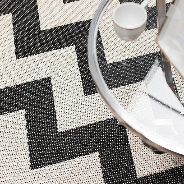 Dekoria Flachgewebeteppich - schwarz, beige, 230 x 160 cm - Nahaufnahme eines modernen Teppichs mit Zickzackmuster in Schwarz und Creme unter einem runden Tisch mit einer Tasse.