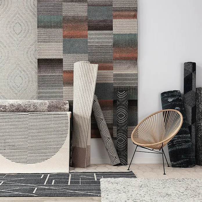 Dekoria Flachgewebeteppich - creme, blau, 290 x 200 cm - Verschiedene Teppiche in verschiedenen Mustern und Farben sind in einem Raum ausgestellt, darunter ein Sessel aus Rattan mit dunklen Metallbeinen.