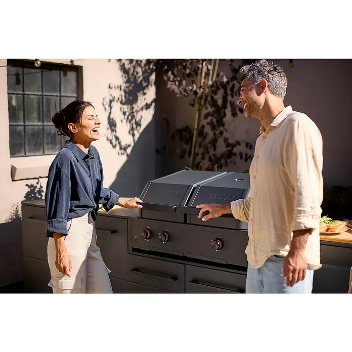 Cozze Hybrid-Grill Quattro 900 - Grillfläche (B x T): 72,7 x 34,5 cm, 13 kW, Anzahl Brenner: 3 Stk. - Lachendes Paar steht vor einer modernen Gozney-Außenküche mit Grill und Arbeitsflächen.