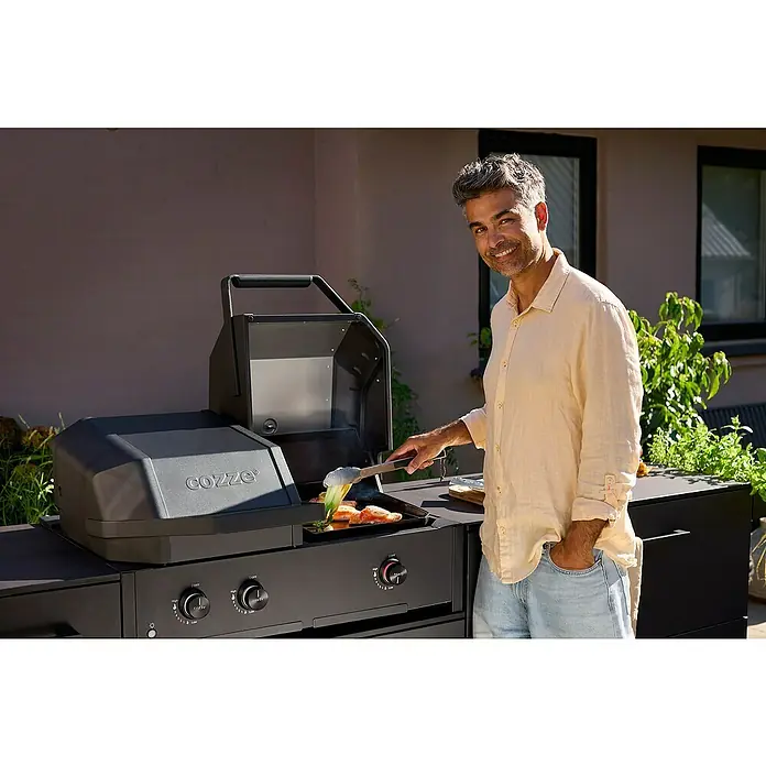 Cozze Hybrid-Grill Quattro 900 - Grillfläche (B x T): 72,7 x 34,5 cm, 13 kW, Anzahl Brenner: 3 Stk. - Ein Mann grillt mit einem Cozze-Grill im Freien, der eine moderne Kochlösung für den Außenbereich bietet.