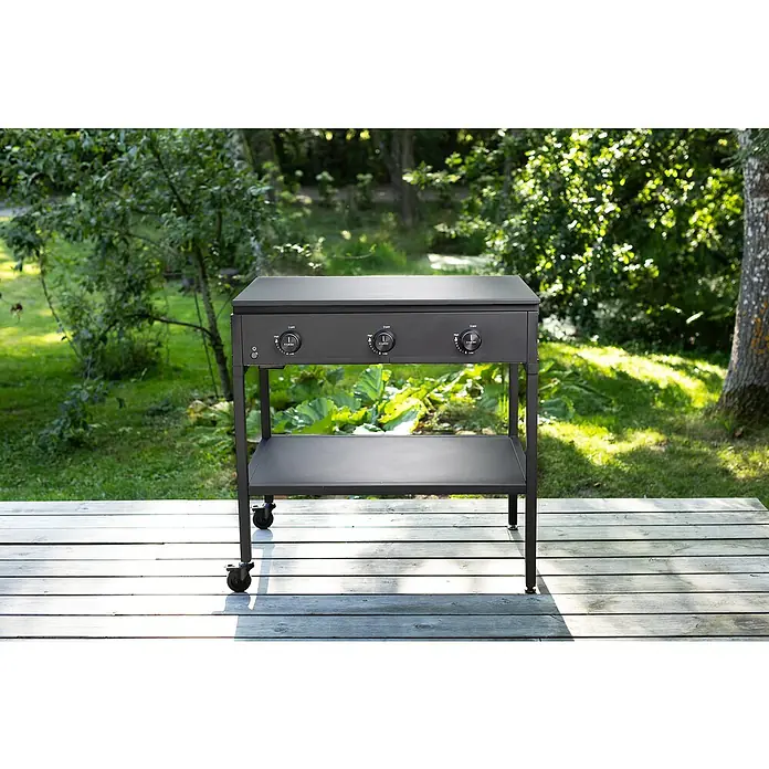 Cozze Grilltisch - B x T x H: 90 x 60 x 71 cm, Stahl, Passend für: Cozze Grills der 900er Serie - Das Bild zeigt einen schwarzen Grillwagen im Freien auf einer Holzterrasse, umgeben von Grün. Der Wagen hat Regler und eine Ablagefläche.