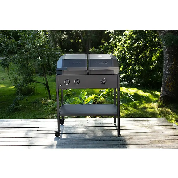 Cozze Grilltisch - B x T x H: 90 x 60 x 71 cm, Stahl, Passend für: Cozze Grills der 900er Serie - Ein grauer Cozze-Pizzaofen auf einem grauen Untergestell mit Rollen, der auf einer hölzernen Terrasse im Grünen steht.