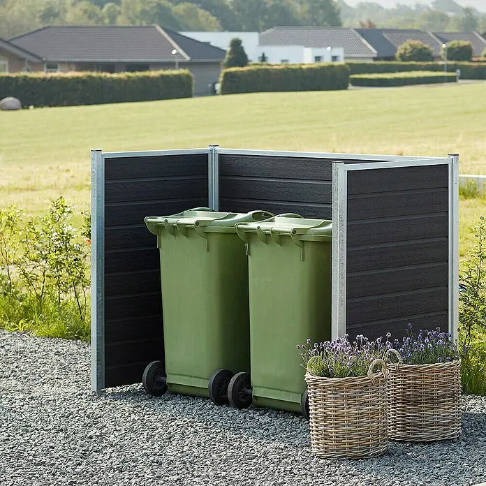 Plus Mülltonnenverkleidung Artura - Holz, Schiefergrau - Das Bild zeigt eine Mülltonnenbox mit zwei grünen Mülltonnen, umgeben von Lavendel in Weidenkörben, vor einem gepflegten Rasen und Häusern.
