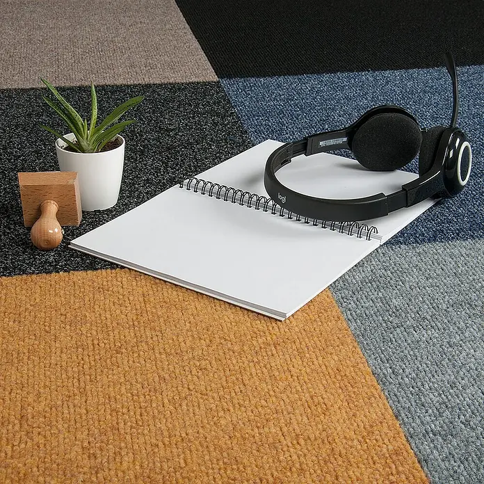 Bricoflor Teppichfliese - Breite: 50 cm, Nadelfilz, Polypropylen, grau - Das Bild zeigt ein Logitech-Headset, das auf einem Notizblock liegt, neben einer Sukkulente und Holzobjekten auf einem bunten Teppich.