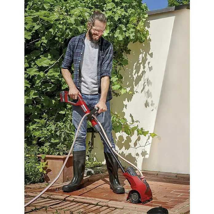 Einhell Power X-Change 18V Akku-Oberflächenbürste Picobella 18/215 - 18 V, Ohne Akku - Ein Mann benutzt eine rote Einhell Fugenbürste mit Wasseranschluss, um Unkraut von einem Gehweg zu entfernen.
