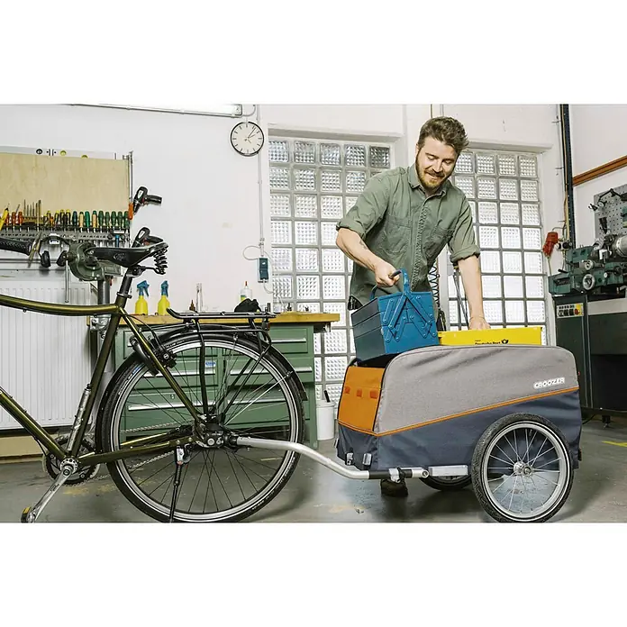 Croozer Fahrradanhänger Cargo Pakko - Traglast: 45 kg, Farbe: Campfire red - Ein Mann belädt einen Croozer Fahrradanhänger mit einer blauen Werkzeugkiste und einem gelben Paket in einer Werkstatt.
