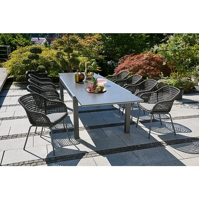 Sunfun Gartenmöbel-Set Maja Stone/Sarah - 9 -tlg., Aluminium, Stahl, Naturstein, Olefin, Night-Grey, Anthrazit - Ein grauer Gartentisch mit acht passenden Rattanstühlen mit grauen Sitzkissen, arrangiert auf einer gepflasterten Terrasse mit üppigem Grün im Hintergrund.