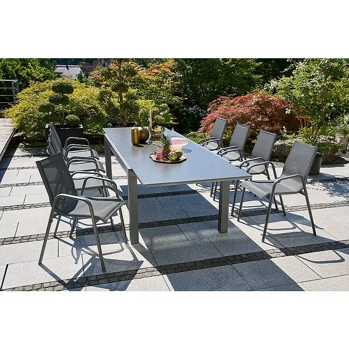 Sunfun Gastro Gartensessel - Stahl, Textilen, Stapelbar, Night-Grey, B x T x H: 57,5 x 68,5 x 88 cm - Ein grauer Gartentisch mit mehreren Stühlen im Freien, umgeben von üppigem Grün. Perfekt für Mahlzeiten im Freien.