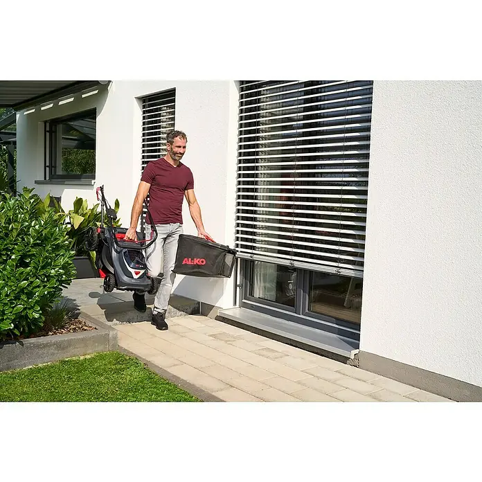 AL-KO 18V/36V Akku-Rasenmäher 43.6 - 18 V - 36 V, 2 Akkus, 5 Ah, Schnittbreite: 43 cm - Ein Mann trägt einen AL-KO Rasenmäher und einen Fangkorb vor einem modernen Haus mit Jalousien.