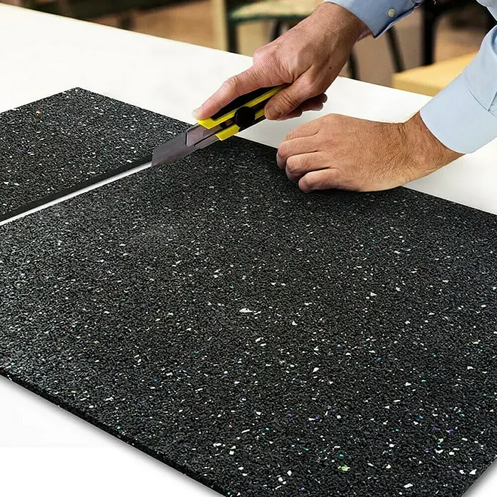 Floordirekt Unterlagsmatte Bautenschutzmatte Gummigranulatmatte 62,5 x 600 x 0,5 cm - Stärke: 5 mm - Eine Person schneidet eine schwarze Gummimatte mit einem gelben Teppichmesser auf einem weißen Tisch. Die Matte besteht aus recyceltem Gummigranulat.