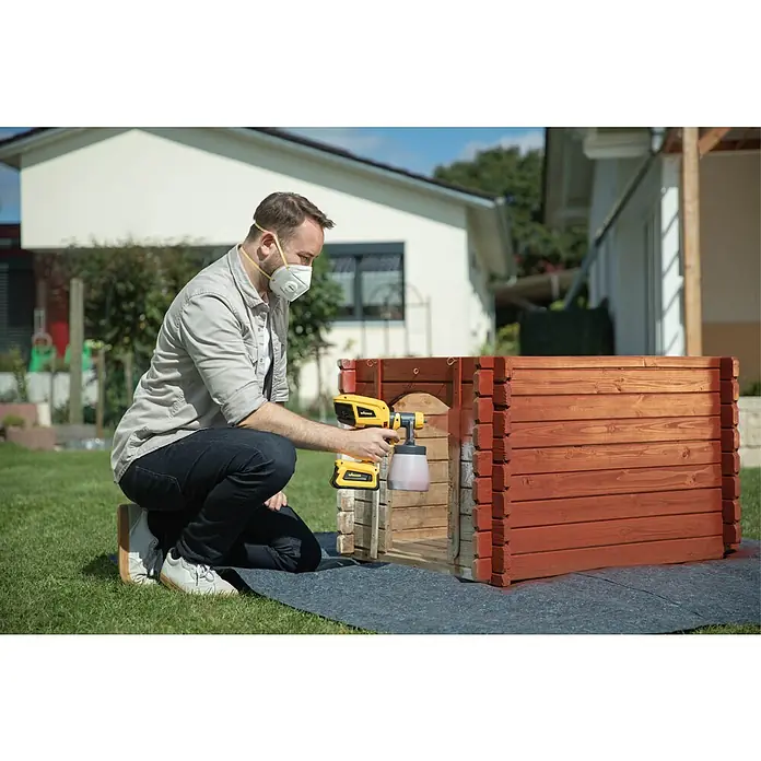 Wagner Akku-Farbsprühsystem W 600 FLEXiO 18V - 18 V, Ohne Akku, Behältervolumen: 1.300 ml, Arbeitsgeschwindigkeit: 2,5 m²/min - Ein Mann lackiert ein Hundehaus mit einer Wagner Farbspritzpistole im Garten, umgeben von Grün. Präzise und effizient.