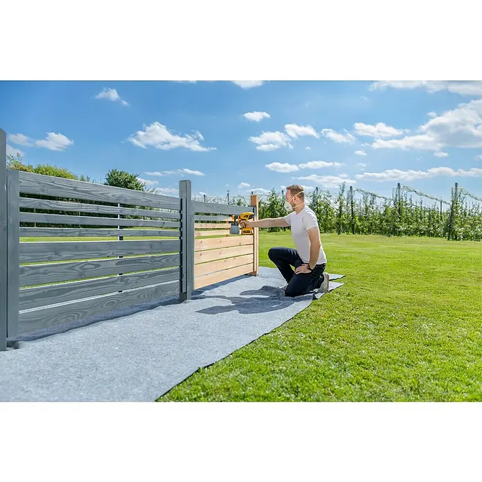 Wagner Akku-Farbsprühsystem W 600 FLEXiO 18V - 18 V, Ohne Akku, Behältervolumen: 1.300 ml, Arbeitsgeschwindigkeit: 2,5 m²/min - Ein Mann streicht einen Holzzaun im Garten mit einer gelben Farbspritzpistole, umgeben von Grün und blauem Himmel. Schutzkleidung ist auch im Einsatz.