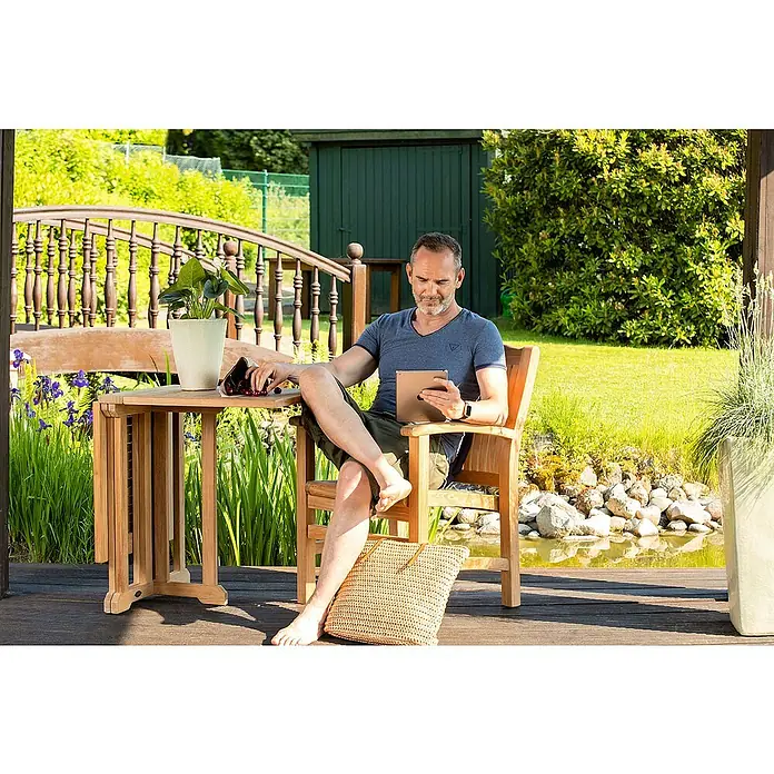 Kai Wiechmann Balkontisch - L x B x H: 120 x 60 x 76 cm, Teakholz, Klappbar, Rechteckig, Naturbelassen - Ein Mann sitzt im Garten auf einem Sessel am Tisch und genießt die Sonne. Der Tisch und Sessel sind aus hochwertigem Teakholz gefertigt.