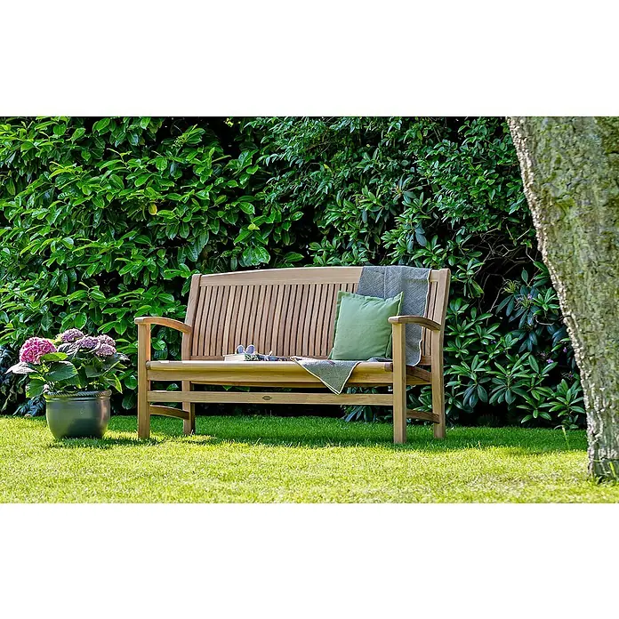 Kai Wiechmann Gartenbank - B x T x H: 200 x 63 x 92 cm, Teak, Naturbelassen - Eine hölzerne Gartenbank mit Kissen und Decke steht auf einem Rasen vor grünen Büschen und einem Baumstamm. Ein Blumentopf ergänzt das Bild.