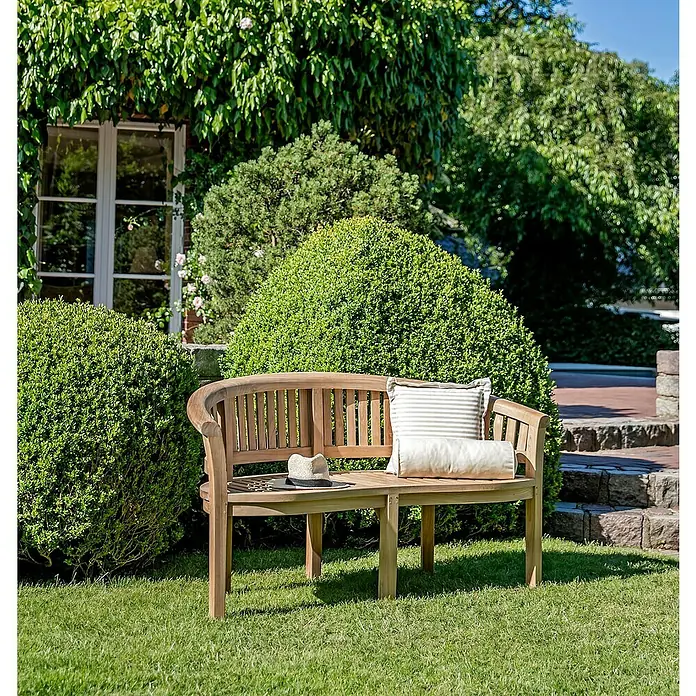 Kai Wiechmann Gartenbank - Teak, Naturbelassen - Eine Gartenbank aus Holz mit Kissen und Hut im Grünen, umgeben von Büschen. Ideal zum Entspannen im Garten.