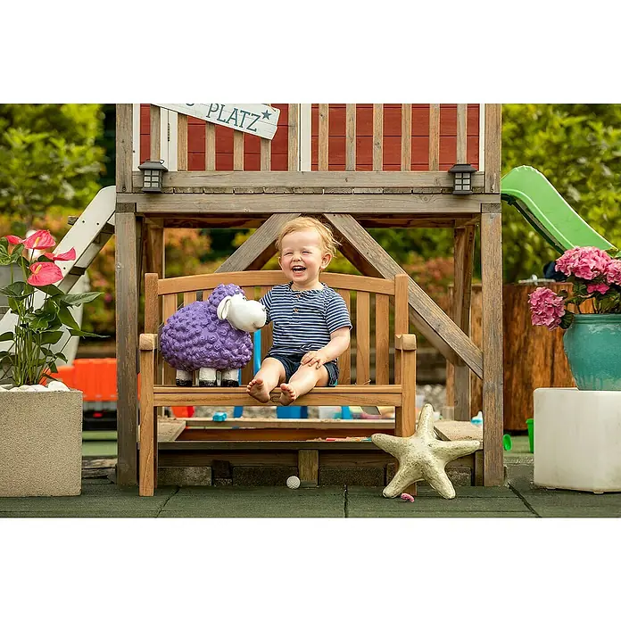 Kai Wiechmann Kinder-Gartenstuhl - Teakholz, Naturbelassen, B x T x H: 80 x 44 x 66 cm - Lachendes Kleinkind sitzt auf einer Gartenbank neben einem lila Schaf. Im Hintergrund befindet sich ein Spielplatz.