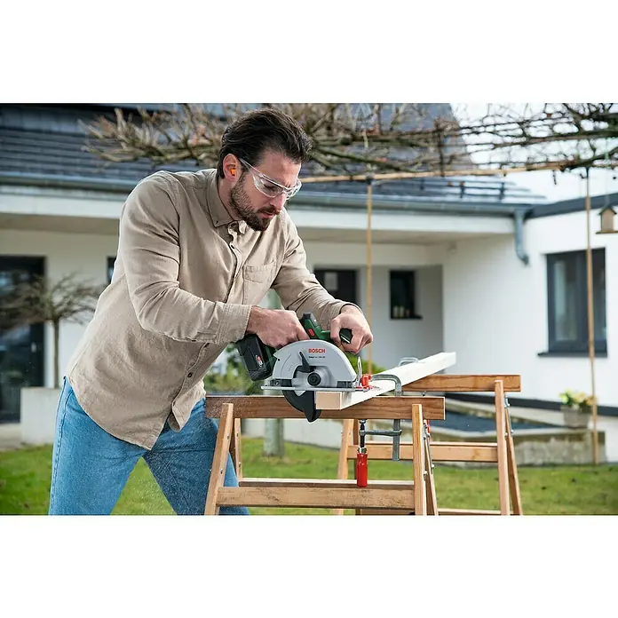 Bosch Power for All 18V Akku-Handkreissäge EasyCirc 18V-48 - 18 V, Ohne Akku, Durchmesser Sägeblatt: 150 mm, 4.200 U/min - Ein Mann sägt ein Holzbrett mit einer Bosch Akku-Kreissäge im Freien, während er Schutzbrille und Ohrenstöpsel trägt.