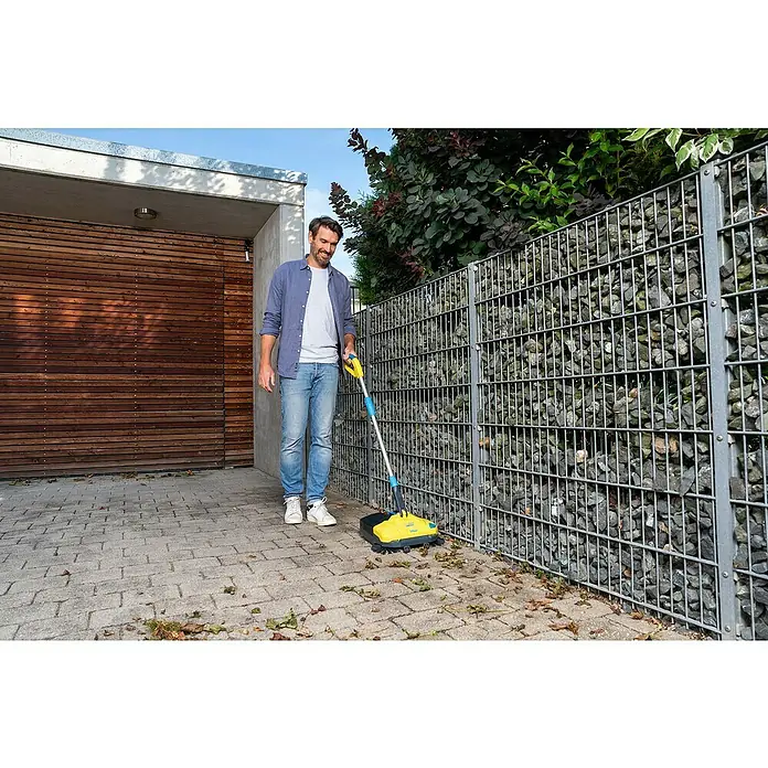Gloria Power for All 18V Akku-Handkehrmaschine EasySweep 18V - 18 V, 1 Akku, Arbeitsbreite: 40 cm, 4,5 l - Ein Mann benutzt eine gelbe Kehrmaschine, um Blätter auf einer gepflasterten Fläche neben einem Haus und einem Steingarten zu entfernen.