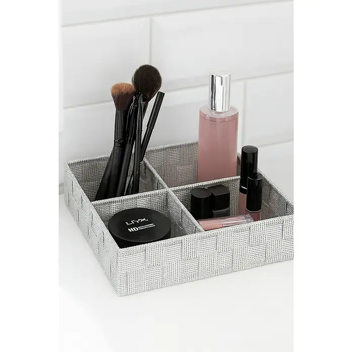 Versa Cesta para el baño 4 - L x An x Al: 32 x 27 x 10 cm, Blanco - Un organizador de cosméticos gris tejido con varios compartimentos, que contiene pinceles, maquillaje y lociones, sobre una encimera blanca.