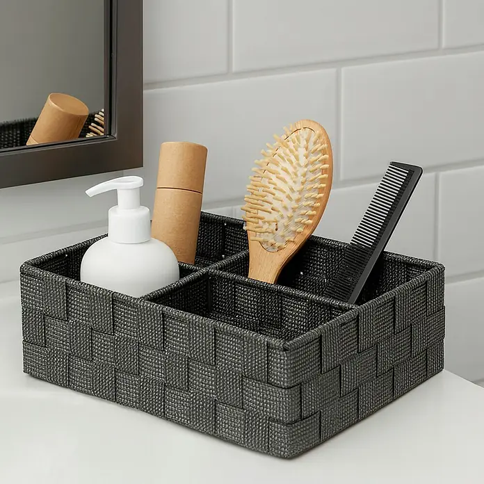 Versa Cesta para el baño 4 - L x An x Al: 32 x 27 x 10 cm, Gris - Un organizador de baño gris tejido lleno de varios artículos de baño, incluyendo un dispensador de jabón, un cepillo de pelo y un peine.