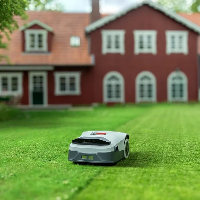 Anthbot Mähroboter Genie 3000 - 3.600 m², Schnittbreite: 20 cm, Satellitengestützte Navigation, RTK - Ein Rasenmähroboter auf einem grünen Rasen mit einem roten Haus im Hintergrund. Der Rasenmähroboter ist grau und weiß.