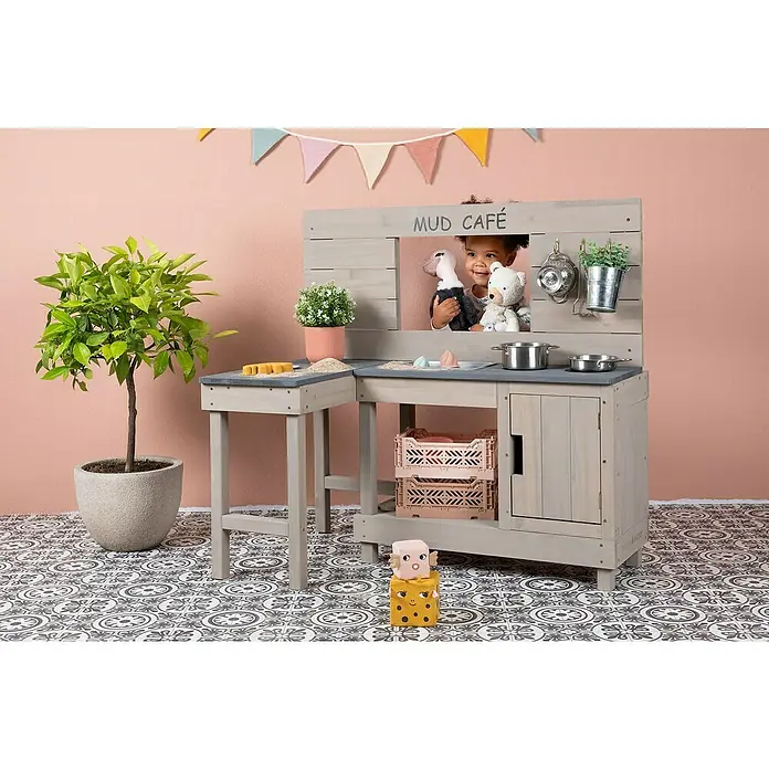 Muddy Buddy Matschküche Coffee Corner - 100 x 60 x 95 cm, Holz, Warmgrau - Ein grauer Spielküche mit "Mud Café" Aufschrift, Töpfen und Pflanzen, dekoriert mit einer bunten Wimpelkette, steht auf einem gemusterten Boden.