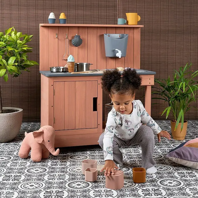 Muddy Buddy Matschküche Mud Expert - 80 x 40 x 90 cm, Holz, Puderrosa - Ein detailreiches Bild einer Spielküche aus Holz mit Zubehör, umgeben von Pflanzen und einem Kind, das mit Spielzeug spielt.