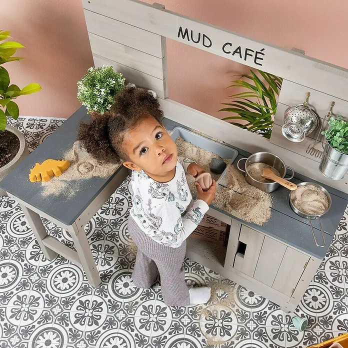 Muddy Buddy Matschküche Coffee Corner - 100 x 60 x 95 cm, Holz, Warmgrau - Ein kleines Mädchen spielt mit einer Mud-Café-Spielküche, die mit Zubehör wie Töpfen und einer Form für Sand ausgestattet ist.