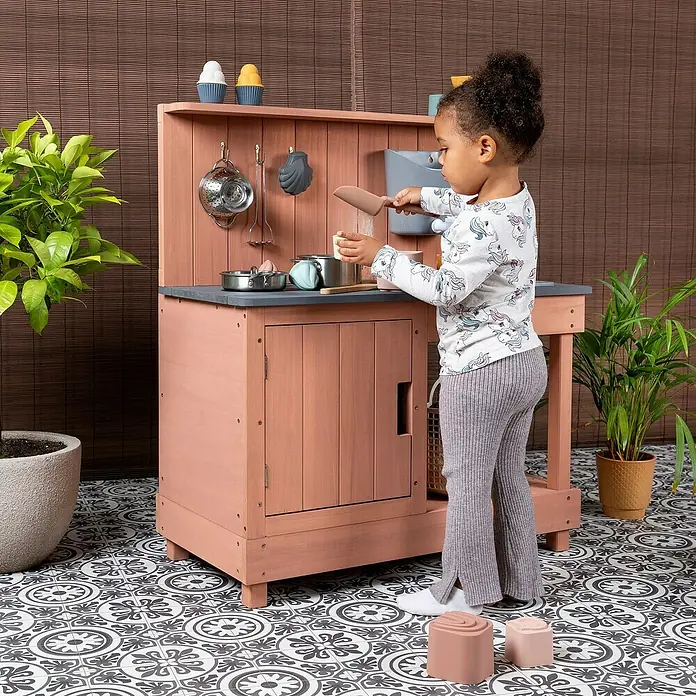Muddy Buddy Matschküche Mud Expert - 80 x 40 x 90 cm, Holz, Puderrosa - Ein Kind spielt mit einer Outdoor-Spielküche aus Holz mit Zubehör, um fantasievolles Spielen im Freien zu fördern.
