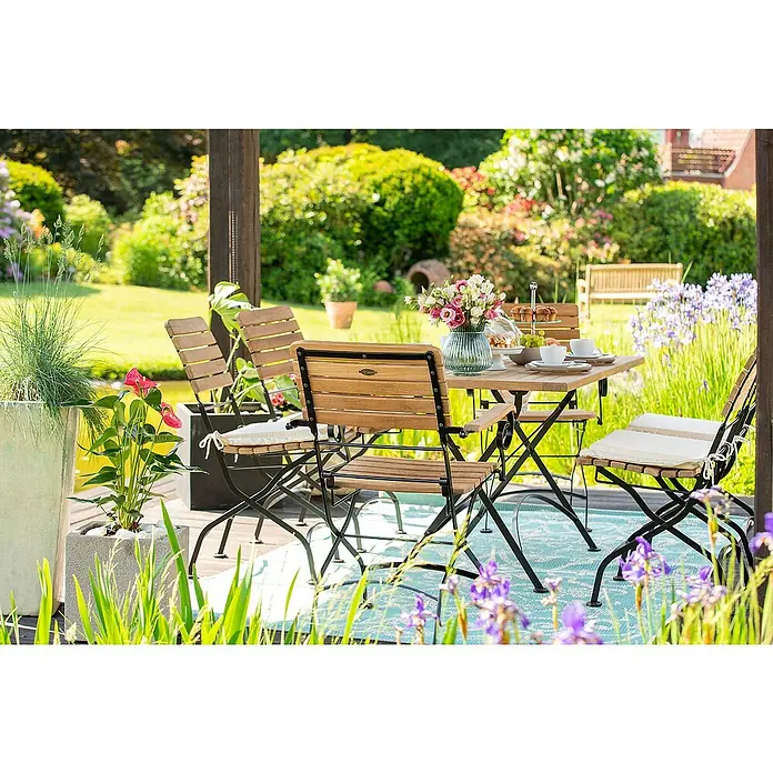 Kai Wiechmann Gartenstuhl - Teakholz, Klappbar, Naturbelassen, B x T x H: 45 x 60 x 89 cm - Eine einladende Gartenszene mit einem eleganten Holztisch und Stühlen, perfekt für Mahlzeiten im Freien inmitten blühender Pflanzen.