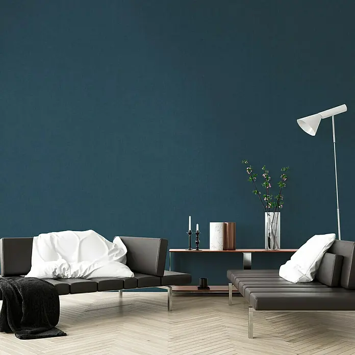 A.S. Création New Walls Vliestapete Textil - Blau/Grün, Uni, 10,05 x 0,53 m - Das Bild zeigt ein modernes Wohnzimmer mit einem grauen Sofa, weißen Kissen und einer minimalistischen Lampe vor einer tiefblauen Wand.