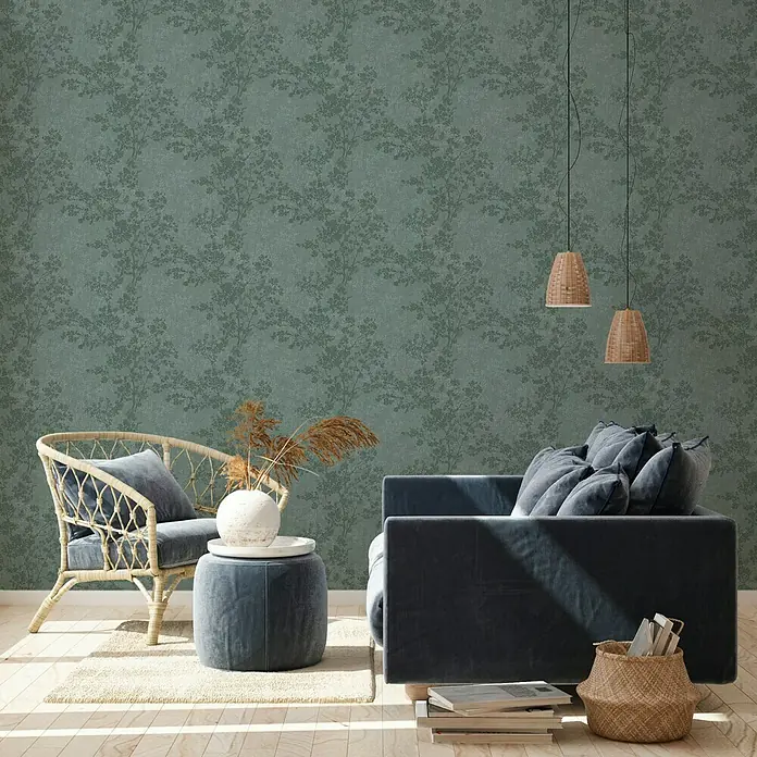 A.S. Création New Walls Vliestapete Bäume - Dunkelgrün, Floral, 10,05 x 0,53 m - Ein stilvolles Wohnzimmer mit einem blauen Samtsofa, einem Rattan-Sessel und einer dekorativen Tapete mit Pflanzenmuster.