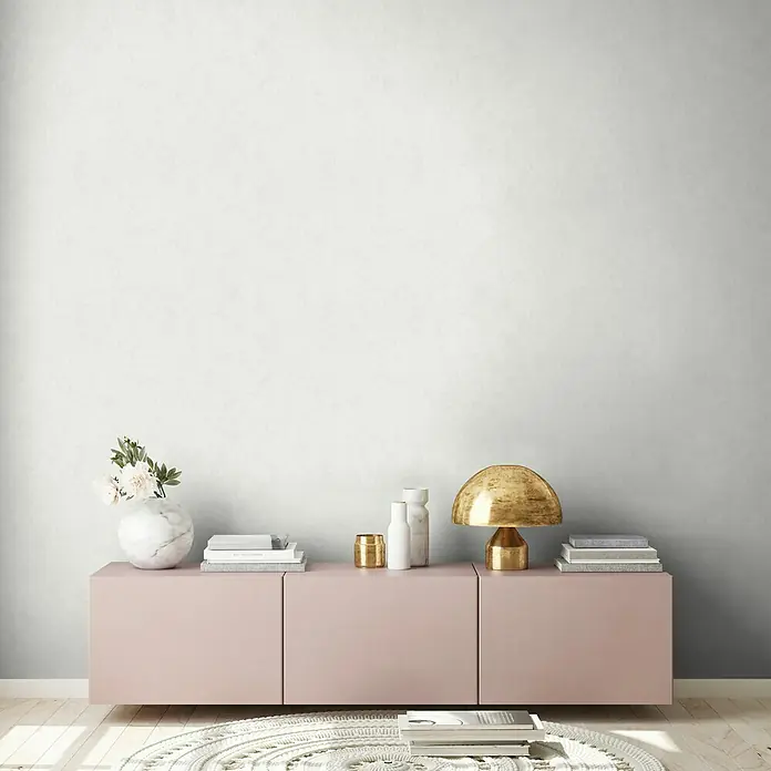 A.S. Création New Walls Vliestapete Textil - Hellgrau, Uni, 10,05 x 0,53 m - Eine moderne rosa Kommode mit dekorativen Elementen wie einer goldenen Lampe, Büchern und einer Vase mit Blumen.
