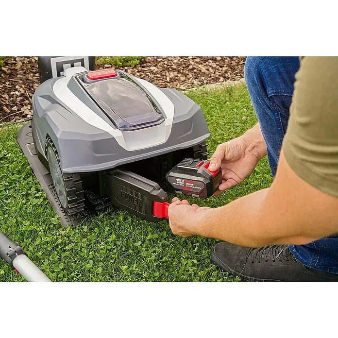 AL-KO 18V/36V Mähroboter Robolinho 1000 Vision - 1.000 m², Schnittbreite: 21 cm, Kamera-gestützte Navigation - Ein Mann setzt einen 18V Akku in einen grauen Rasenmähroboter ein. Der Akku ist rot und schwarz, und der Rasenmäher steht auf grünem Gras.