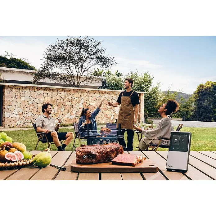 Typhur Grill-Thermometer Sync Dual - 3 -tlg., Kabellos; Verbindung via Bluetooth oder Wi-Fi - Gruppenfoto mit dem Typhor Fleischthermometer auf einem Holztisch mit Früchten und gegrilltem Fleisch, während Leute grillen.