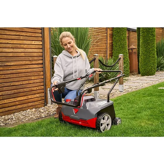 AL-KO 18V/36V Akku-Vertikutierer 38.6 - Ohne Akku, Arbeitsbreite: 38 cm - Frau bedient einen Rasenlüfter im Garten. Der Rasenlüfter ist rot-grau und steht auf einem grünen Rasen.