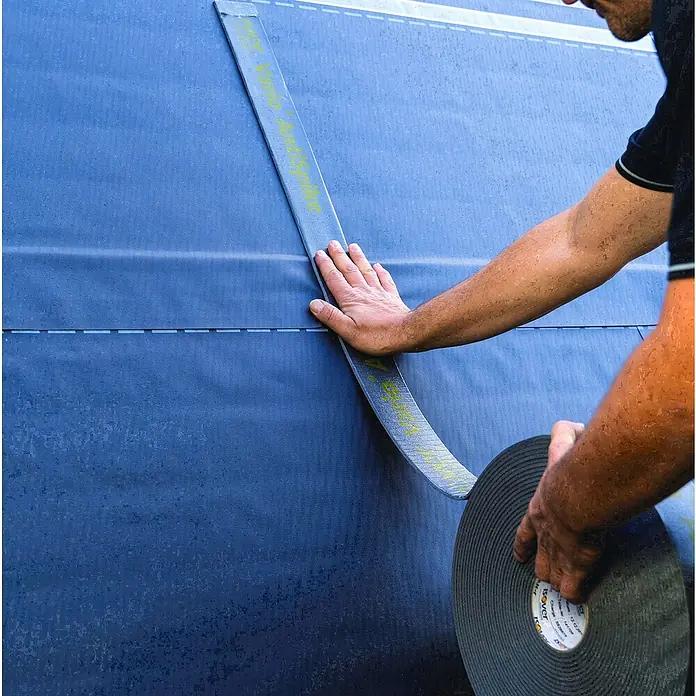Isover Vario Nageldichtband AntiSpike - B x L: 65 mm x 20 m - Eine Person befestigt ein Dichtungsband von Isover an einer Wand, um diese vor Feuchtigkeit zu schützen. Das schwarze Band wird sorgfältig angebracht.