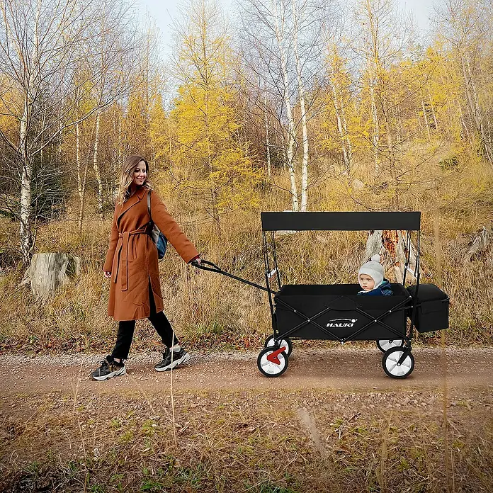 Hauki Bollerwagen - L x B x H: 170 x 53 x 104 cm, Stahl - Eine Frau zieht einen schwarzen Hauck Bollerwagen mit einem Kind darin auf einem herbstlichen Weg.
