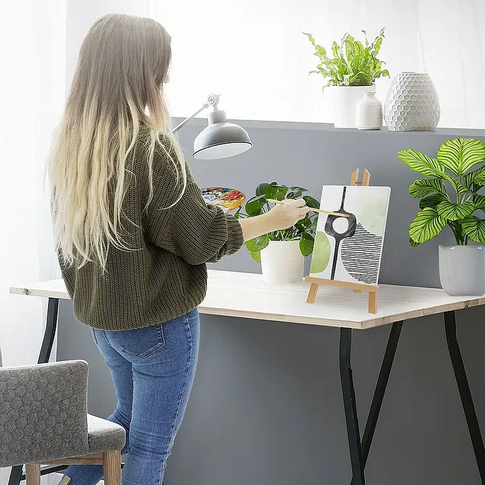 ECD Germany Zeichenstift-Set Tischstaffelei aus Holz klappbar mit H-Rahmen - Braun - Eine Frau mit einem Pinsel in der Hand steht vor einer Leinwand, die auf einer Staffelei steht, und malt ein abstraktes Kunstwerk.