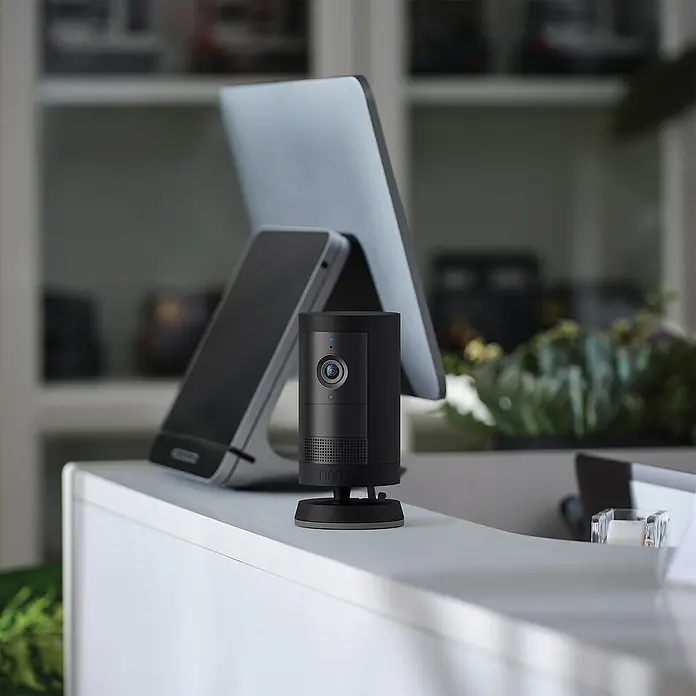 Ring Außenkamera Plus (Plug) - 2.048 x 1.536 Pixel (2K-HD), Schwarz - Das Bild zeigt eine Ring Sicherheitskamera, die auf einem weißen Tisch vor einem Tablet und einem Bücherregal platziert ist.